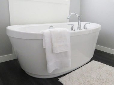 An inviting modern white free standing bathtub in a remodeled bathroom with a wood floor and bathmat